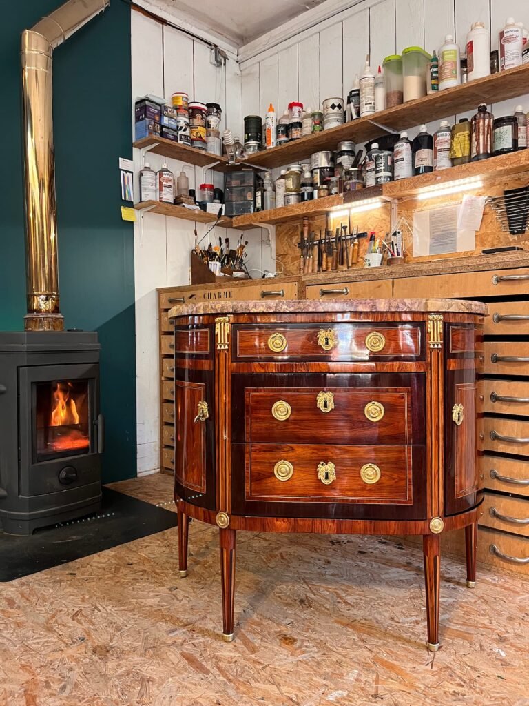 Commode ancienne restaurée exposée dans un atelier d’ébénisterie artisanale avec poêle à bois et outils traditionnels